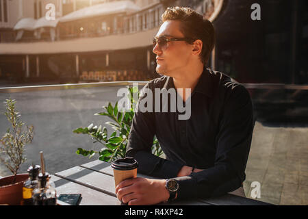 Attraktiven jungen Mann am Tisch und schaut nach links sitzen. Er halten Tasse trinken. Er sitzt draußen. Es ist sonnig. Stockfoto