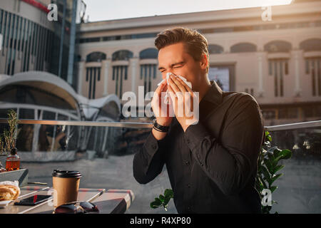 Sixk junger Mann am Tisch sitzen und Niesen in Weiß Serviette. Er hält die Augen geschlossen. Kerl leiden. Er sitzt draußen. Es ist sonnig. Stockfoto