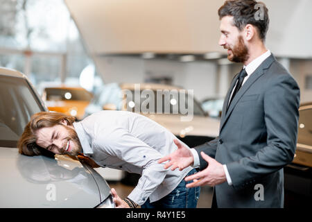 Junger Mann client Berühren mit Gesicht Auto Motorhaube eines neuen Luxus Auto stehend mit Verkäufer im Showroom Stockfoto