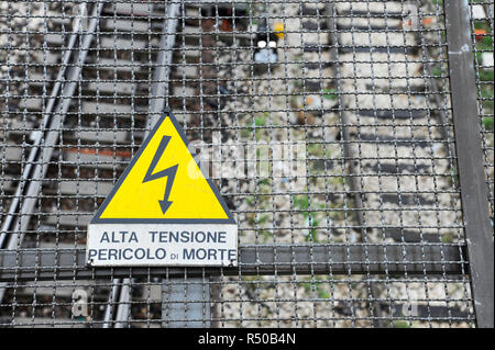 Gelbes Warnschild für Hochspannung Fahrdrähten über den Schienen, im Hintergrund der Bahnhof Tracks. Warnsymbol angezeigt. Bologna, Italien Stockfoto