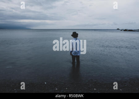 Voll bekleidet Mann mit Hut steht in seichtem Wasser, mit dem Rücken zum Betrachter, wie er aufs Meer schaut, Langzeitbelichtung, Taiwan. Weiche gefilterten Bilder Stockfoto