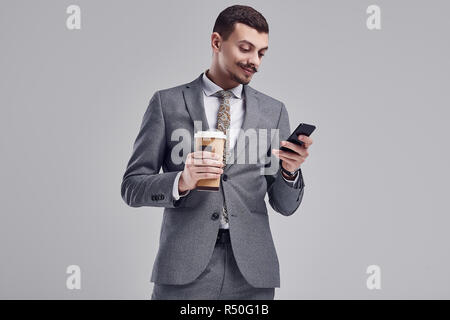 Portrait von gutaussehenden Jungen zuversichtlich arabischer Geschäftsmann mit ausgefallenen Schnurrbart in Mode grauen kompletten Anzug hält eine Tasse Kaffee und eine Meldung eingeben auf Th Stockfoto