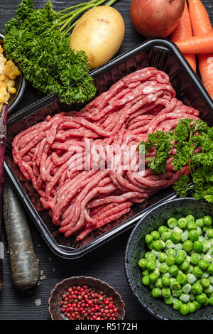 Rohes Hackfleisch in Container mit Zutaten für Shepherds Pie mit grünen Erbsen, gelber Mais, Karotten, Kartoffeln, Petersilie, Zwiebeln und Gewürze, auf Schwarz Stockfoto