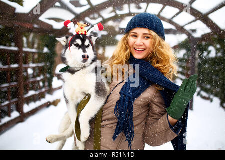 Attraktive authentische kaukasische Frau umarmt Lustig malamute Hund santa tragen liebe Weihnachten Geweih. Curly lächelnd weibliche Spaß mit huskie Welpen im neuen Jahr. Pet ist das beste Geschenk für Feiertage. Stockfoto