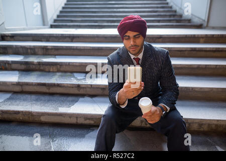 Indischer Geschäftsmann sitzt auf der Treppe, während Sie Telefon und Kaffee trinken Stockfoto