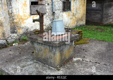 Beton mit Metallabdeckung und grau Eimer mit einem Vorhängeschloss gesichert verrostete Neben Alte kaputte Wasserpumpe vor der abgebrochenen alten verfallenen Haus Stockfoto