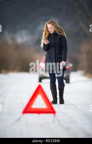 Junge Frau ein Warndreieck Einrichtung und Unterstützung gefordert, nachdem ihr Auto in der Mitte des nirgendwo an einem eiskalten Wintertag ist kaputt Stockfoto