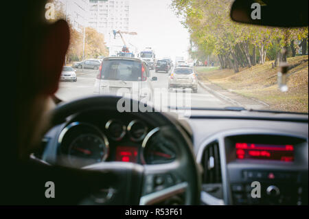 Man fährt ein Auto. Ein Kerl in einer Jacke sitzt hinter dem Steuer eines Autos fahren auf der Straße im Stau. Foto aus dem Sitz, durch Rücken des Fahrers Stockfoto