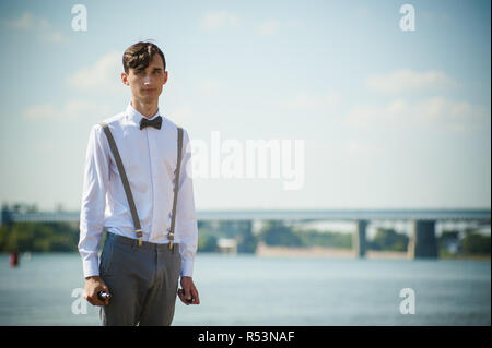 Junge dünne Mann, elegant und stilvoll in weißem Hemd, Hose mit Hosenträgern und Bow Tie. vaper Kerl raucht eine e-Zigarette, releas Rauchschwaden, gegen den Hintergrund von Fluss und Brücke Stockfoto