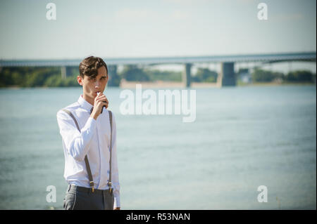 Junge dünne Mann, elegant und stilvoll in weißem Hemd, Hose mit Hosenträgern und Bow Tie. vaper Kerl raucht eine e-Zigarette, releas Rauchschwaden, gegen den Hintergrund von Fluss und Brücke Stockfoto