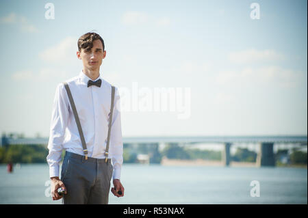 Junge dünne Mann, elegant und stilvoll in weißem Hemd, Hose mit Hosenträgern und Bow Tie. vaper Kerl raucht eine e-Zigarette, releas Rauchschwaden, gegen den Hintergrund von Fluss und Brücke Stockfoto