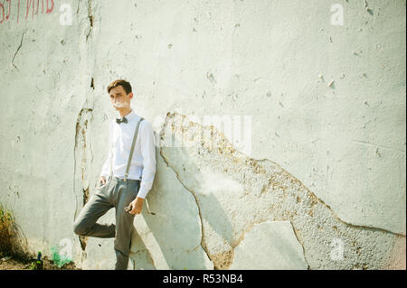 Junge dünne Mann, elegant und stilvoll in weißem Hemd, Grau Hose mit Hosenträgern und Fliege. Raucht eine elektronische Zigarette auf dem Hintergrund von schäbig Wand, warmen, sonnigen Sommer Tag Stockfoto