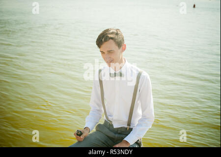 Junge dünne Mann, elegant und stilvoll in weißem Hemd, Grau Hose mit Hosenträgern und Bow Tie. Kerl eine elektronische Zigarette raucht, Rauch, sitzt auf der Pier, gegen den Hintergrund der Fluss Stockfoto