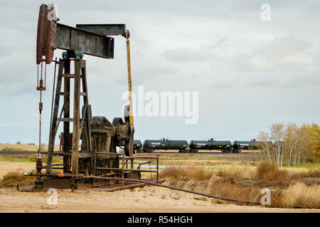 Ein Pumpenheber pumpt Öl aus einem Schieferölbrunnen außerhalb von Williston, einem Abschnitt des Bakken-Ölfeldes. Im Hintergrund ein Güterzug mit Tankwagen voller Öl. Stockfoto