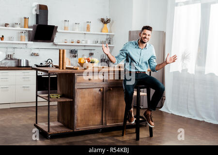 Fröhlicher junger Mann lächelnd in die Kamera beim Sitzen mit offenen Armen in der Küche Stockfoto