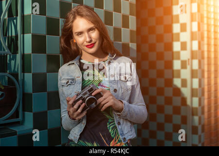 Cool girl Modell mit retro Film Kamera trägt eine Jeansjacke, dunkle Haare im Freien über Stadtmauer in einem Käfig Hintergrund. Spaß in der Stadt mit Cam Stockfoto