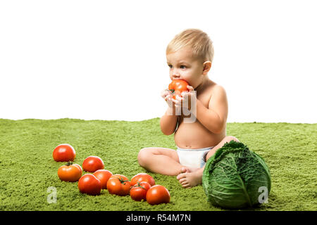 Cute Baby Boy essen Tomaten und sitzen auf grünen Teppich in der Nähe von mit Gemüse isoliert auf weißem Stockfoto