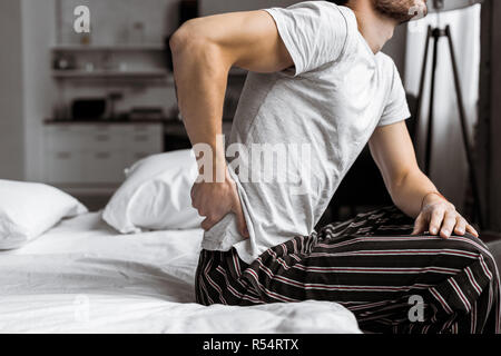 7/8 shot junger Mann im Schlafanzug leiden unter Rückenschmerzen, während auf dem Bett am Morgen sitzen Stockfoto