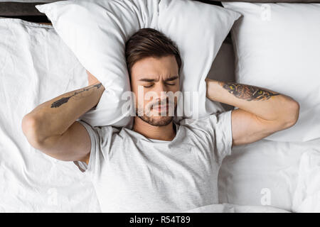 Junger Mann Ohren schließen mit Kissen im Bett am Morgen Stockfoto