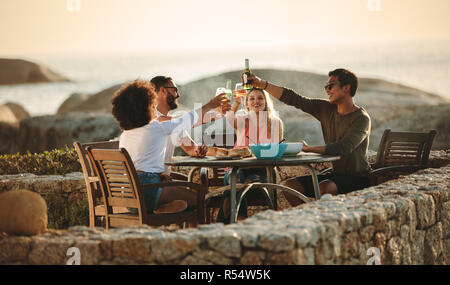Vier Freunde toasten Getränke sitzen auf einem Esstisch im Freien in der Nähe der Küste. Multiethnischen Freunde im Urlaub Spaß haben, Wein trinken und Snacks Stockfoto