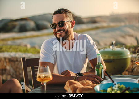 Nahaufnahme von einem lächelnden Mann in Sonnenbrille an einem Tisch mit Speisen und Getränken sitzen. Mann, der an einem Urlaub in ländlicher Umgebung Spaß mit Freunden ea Stockfoto