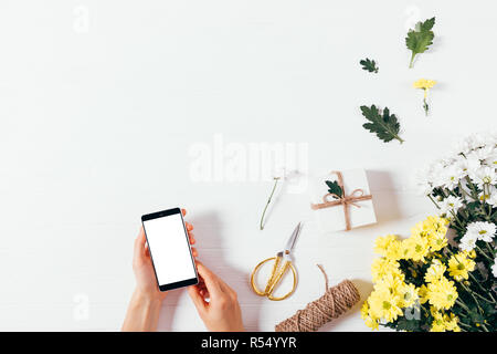 Woman's Hände, die smart phone mit leeren Bildschirm auf weißen Tisch in der Nähe von dekorativen flach Zusammensetzung der gelben Blumen und Geschenk Box, Ansicht von abov Stockfoto