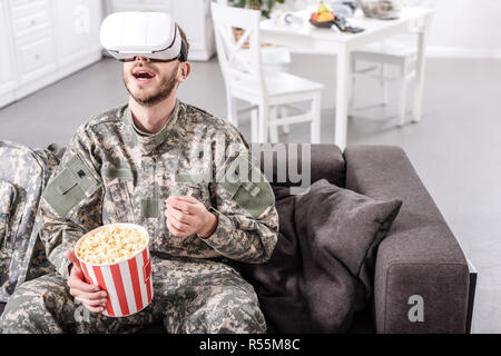 Aufgeregt Soldat in virtual reality Headset sitzen auf der Couch und essen Popcorn Stockfoto