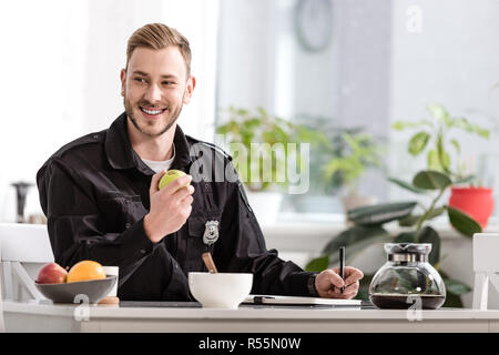 Lächelnd Polizisten schreiben im Notebook und essen Apple am Küchentisch Stockfoto