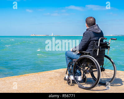 Behinderte junge Mann sitzt im Rollstuhl und schaut auf das Meer Stockfoto