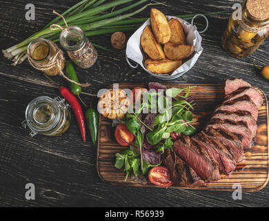 Geschnittenes saftiges Steak medium auf der Platine mit Kräutern, Bratkartoffeln, Gewürze auf einem schwarzen Oberfläche Stockfoto