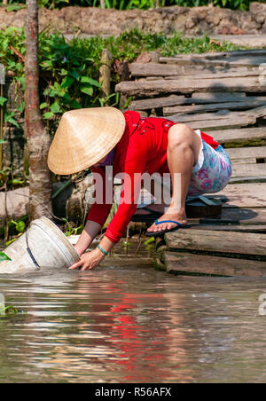 Junge Frau mit leuchtend roten Top zusammengekauert am Wasser aus einem Eimer in Fluss in der Provinz Can Tho, Vietnam zu waschen. Stockfoto