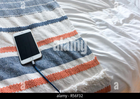 Ein iphone an eine Schnur, ruht auf einem schönen gewebte Decke auf einem luxuriösen Bett suchen. Zimmer für Kopie Stockfoto