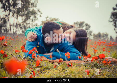 Junge Paare, die auf dem Gras in einem Feld von roter Mohn und bei jedem anderen lächelnd Stockfoto