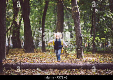 Das Thema Kinder Aktivitäten im Freien. Lustige kleine Baby Kaukasier blonde Mädchen Spaziergänge durch Wald Hindernisse überwinden, Baum, log. Baby Wandern große lustig Rucksack im Herbst Wald Park Stockfoto