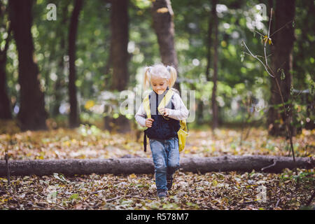 Das Thema Kinder Aktivitäten im Freien. Lustige kleine Baby Kaukasier blonde Mädchen Spaziergänge durch Wald Hindernisse überwinden, Baum, log. Baby Wandern große lustig Rucksack im Herbst Wald Park Stockfoto