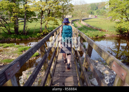 Eine junge weibliche Wanderer entlang eine Fußgängerbrücke über den Fluss Wharfe, in der Nähe von Starbotton, die in den Yorkshire Dales, Northern England, Vereinigtes Königreich. Stockfoto