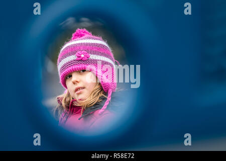 Junge Mädchen in einem rosa Hut spähend durch das Loch zwischen den verschiedenen Verstecke in den Spielplatz Stockfoto