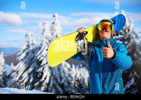 Lächelnd posiert Snowboard Snowboarder Tragen auf den Schultern am Ski Resort in der Nähe von Forest vor Freeride Session. Reiter Daumen oben Zeichen tragen polarisierte Brille. Moderne Snowboardausrüstung. Stockfoto