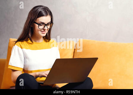 Attraktive Langhaarige brünette Mädchen mit Brille sitzen auf dem gelben Sofa im Wohnzimmer und schreiben ihre Prüfung Test auf dem Laptop, moderne Technik, Kommunikation Konzept Stockfoto