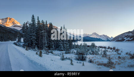 Klein und leer Winter Straße, mit einem kleinen Wald und großen zugefrorenen See bei Sonnenuntergang in wunderschöne Berge, Banff National Park, Kanada Stockfoto