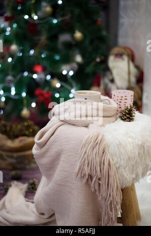 Der Weihnachtszeit. Heiße Schokolade, einer Tasse Cappuccino auf einem Fell Sessel vor einem großen Weihnachtsbaum mit Kugeln und Lichtern. Warmer Schal, Kegel rund um Stockfoto