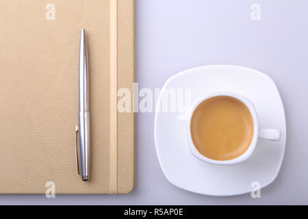 Weiße Tasse mit einem duftenden Espresso, elegantes Notebook und einem Kugelschreiber auf dem Schreibtisch. Ansicht von oben Stockfoto