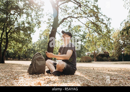 Junge positive Kerl oder Student mit dem Rucksack sitzt und das Hören von Musik oder Podcasts oder Radio in einem Stadtpark. Lebensstil Stockfoto