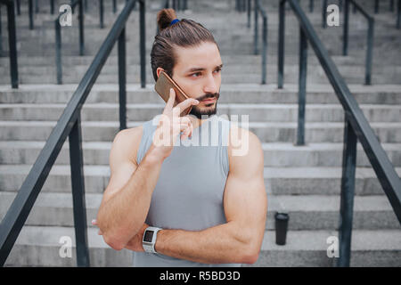 Dunkelhaarige und bärtiger junger Mann steht und Gespräche am Telefon. Er schaut nach rechts. Kerl trägt handband. Es gibt Schritte hinter ihm. Kerl ist allein Stockfoto