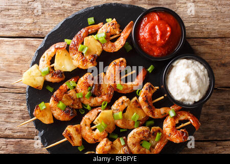 Gegrillte Spieße mit Garnelen mit frischer Ananas und grüne Zwiebeln serviert mit Saucen close-up auf einer Schiefertafel Platte auf einem Holztisch. horizontale Ansicht von oben Stockfoto