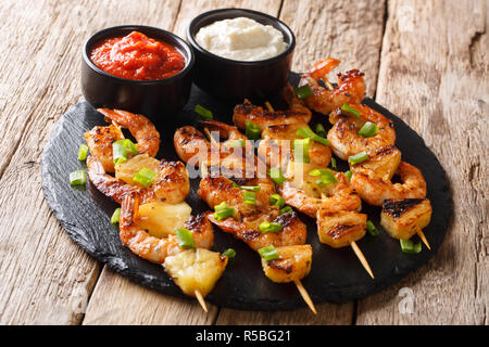 simple homemade recipe for grilled kebabs from shrimp and pineapple slices with sauces close-up on a slate plate on a wooden table. horizontal Stockfoto