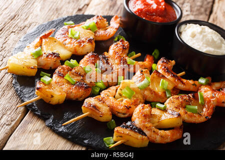 Fresh kebabs made from shrimps and pineapple slices decorated with green onions served with sauces close-up on a slate plate on a wooden table. horizo Stockfoto