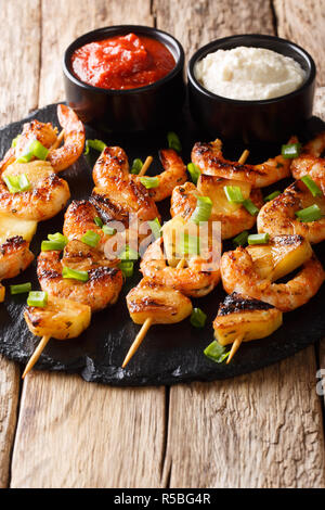 Garnelen kebabs mit Ananas und grüne Zwiebeln mit Soßen schließen eingereicht - bis auf einer Schiefertafel Platte auf einem Holztisch. Vertikale Stockfoto