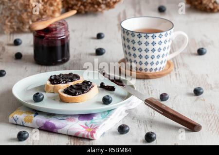 Brot mit Blaubeer-marmelade auf einen Teller, eine Tasse Tee, auf der Rückseite ein Glas von blueberry Jam. Stockfoto