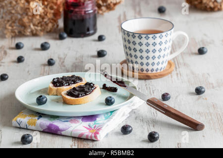 Brot mit Blaubeer-marmelade auf einen Teller, eine Tasse Tee, auf der Rückseite ein Glas von blueberry Jam. Stockfoto